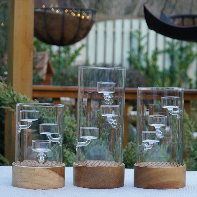 Three glass and wooden cylindrical decorative candle holders on a table with a garden background