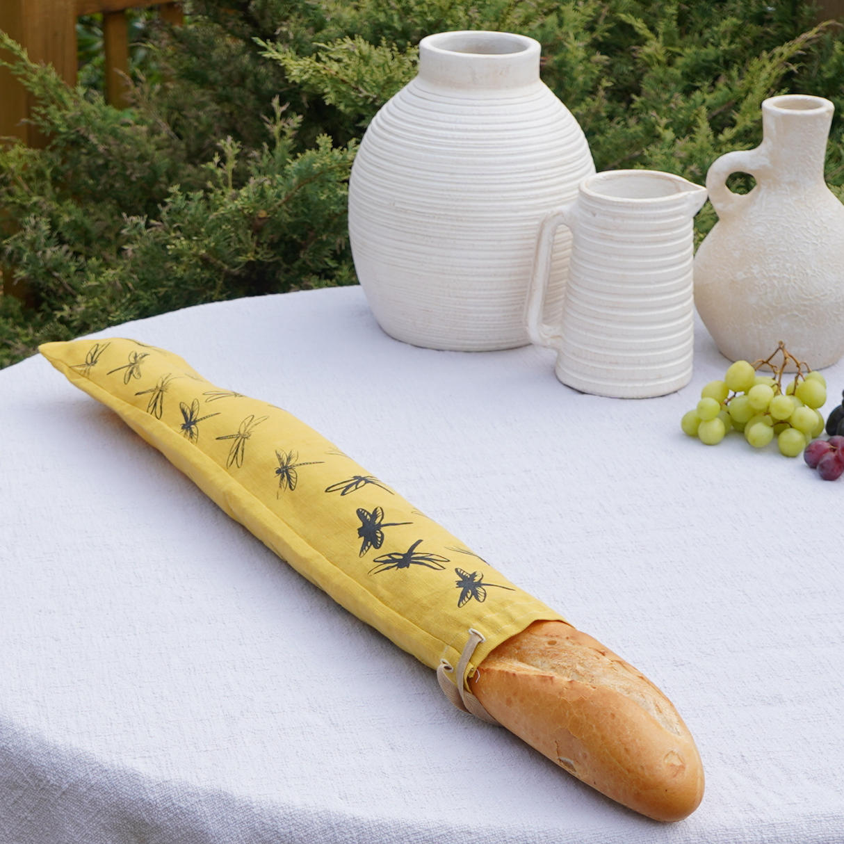 Baguette in a yellow bag with bee pattern on a white tablecloth with ceramic vases and grapes.