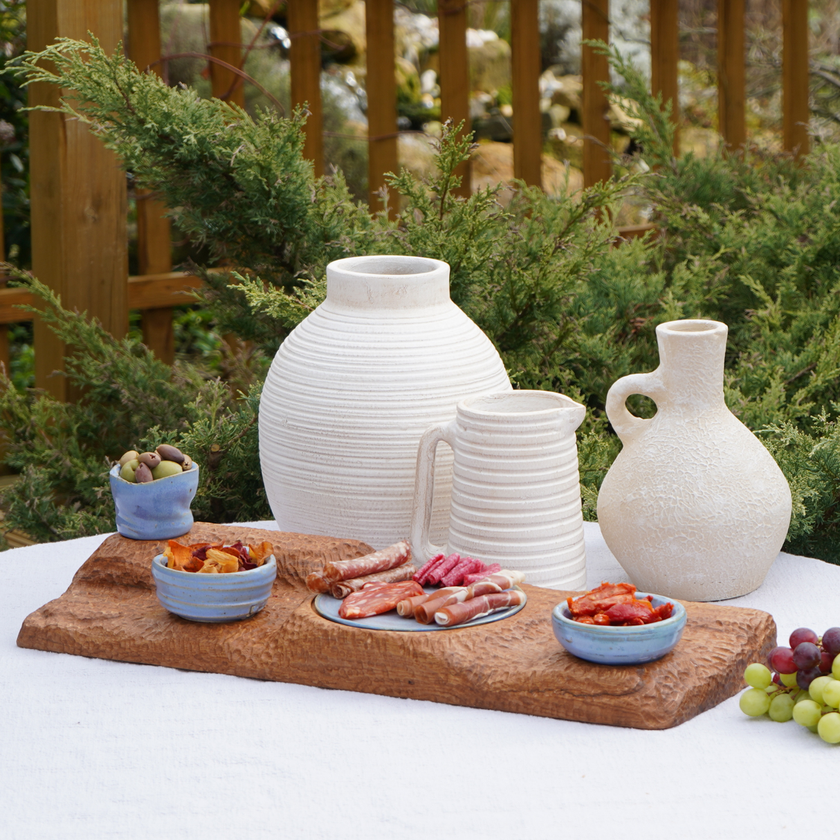 Outdoor setting with ceramic vases and a wooden cutting board with food on a white tablecloth.