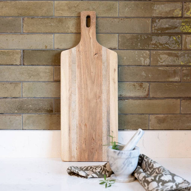 Wooden cutting board leaning against a brick wall with decorative pillows and a plant on a white surface.