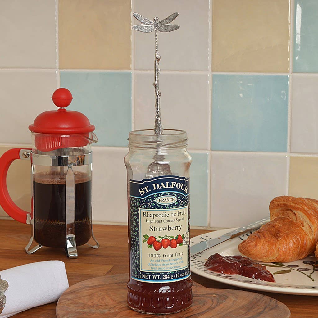 Jar of St. Dalfour strawberry jam with a dragonfly jam spoon on a kitchen counter with coffee and pastries.