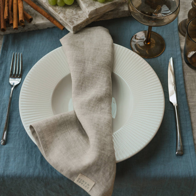 Dining table setting with white plates, gray napkins, silverware, and a blue tablecloth.