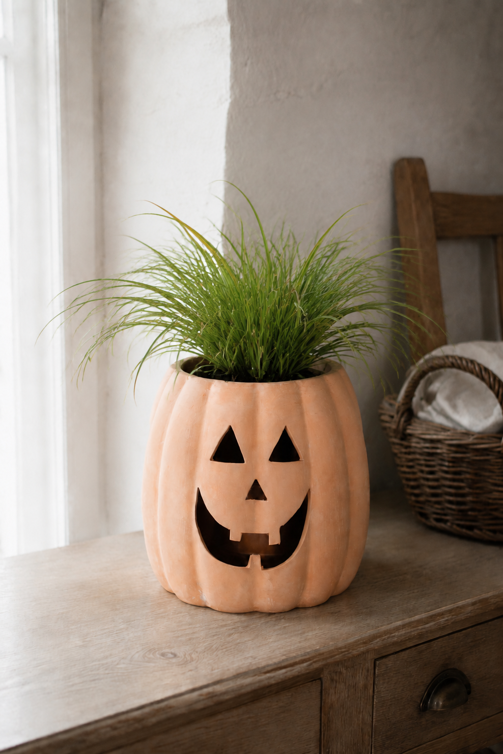 A decorative orange jack-o'-lantern planter with a carved face, filled with green ornamental grass, sitting on a rustic wooden dresser in a softly lit room.