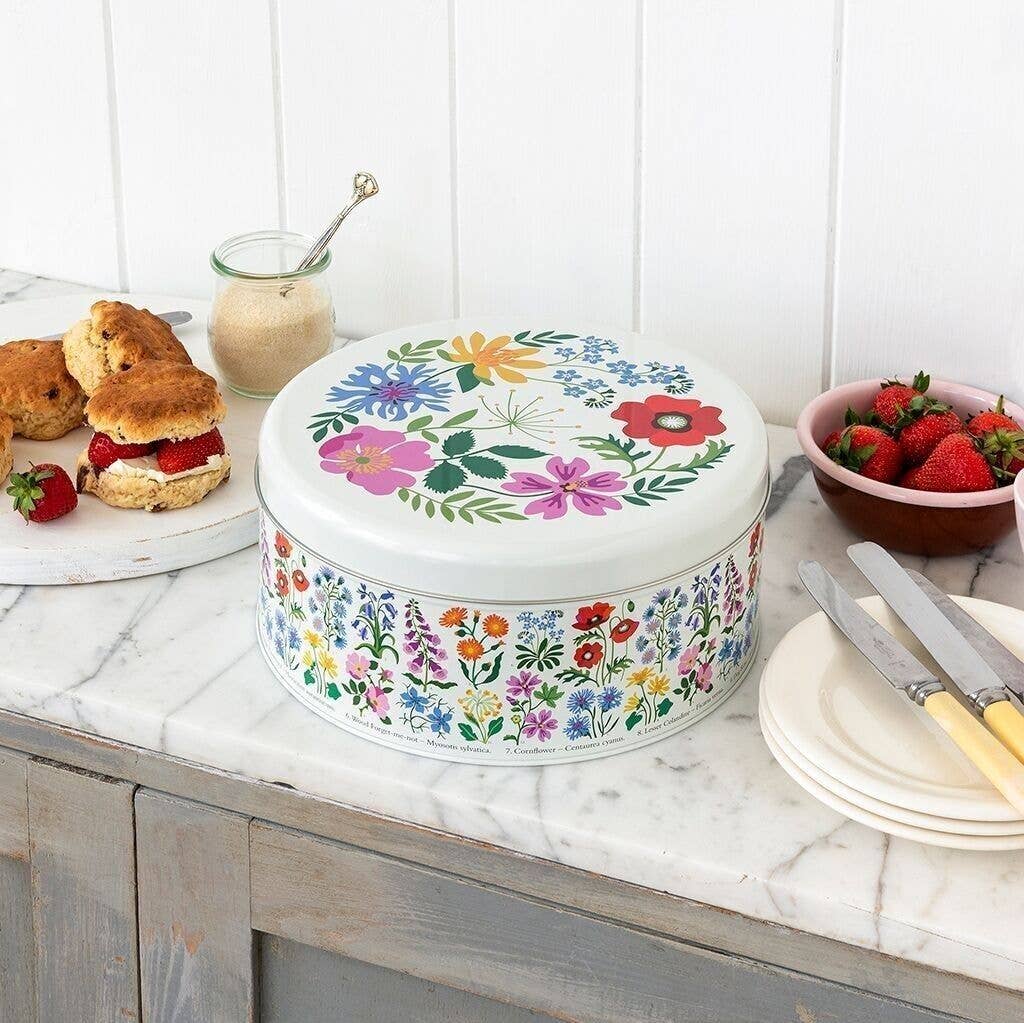A round white tin with a colorful floral print sits on a marble kitchen counter beside scones, strawberries, and plates.