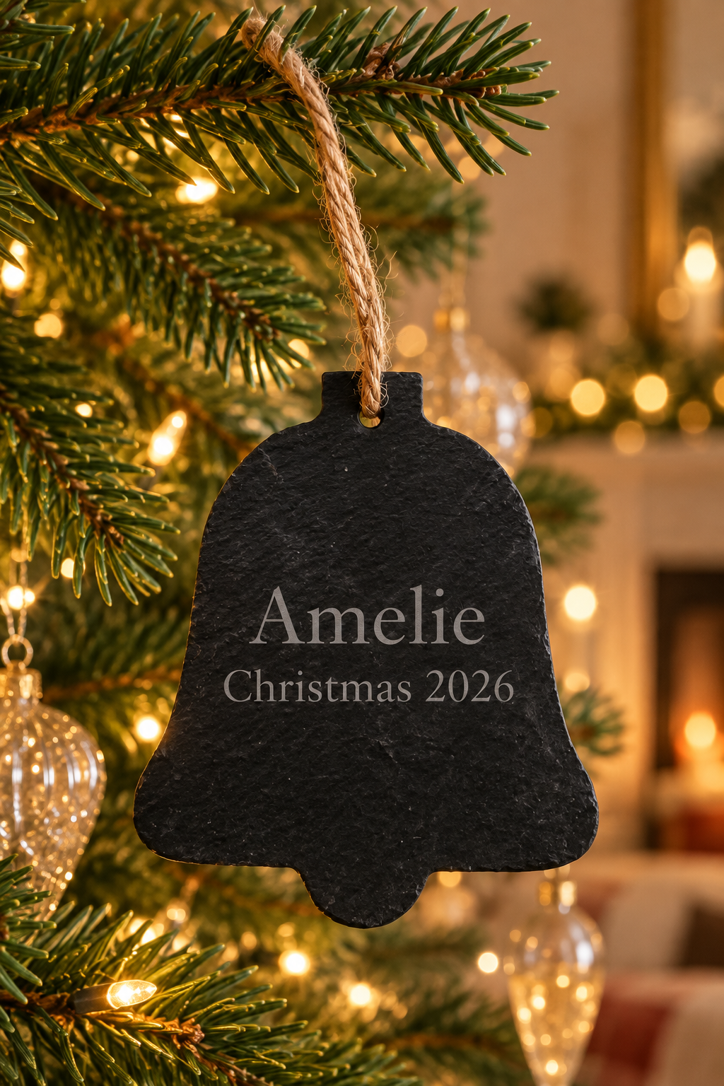 A black slate Christmas bell ornament engraved with 'Amelie Christmas 2026', hanging from a green pine tree branch with warm string lights in the background.