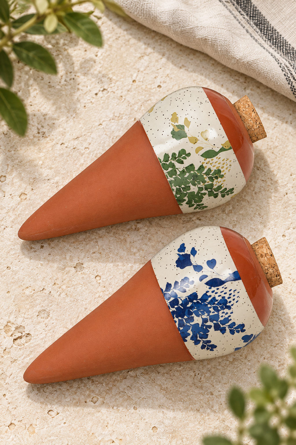 Two Lison terracotta watering spikes with cork stoppers and speckled cream bands, one with blue foliage and one with green and ochre foliage.