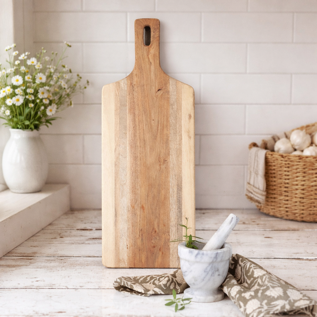 Wooden cutting board on a kitchen counter with a vase of flowers and a basket in the background.