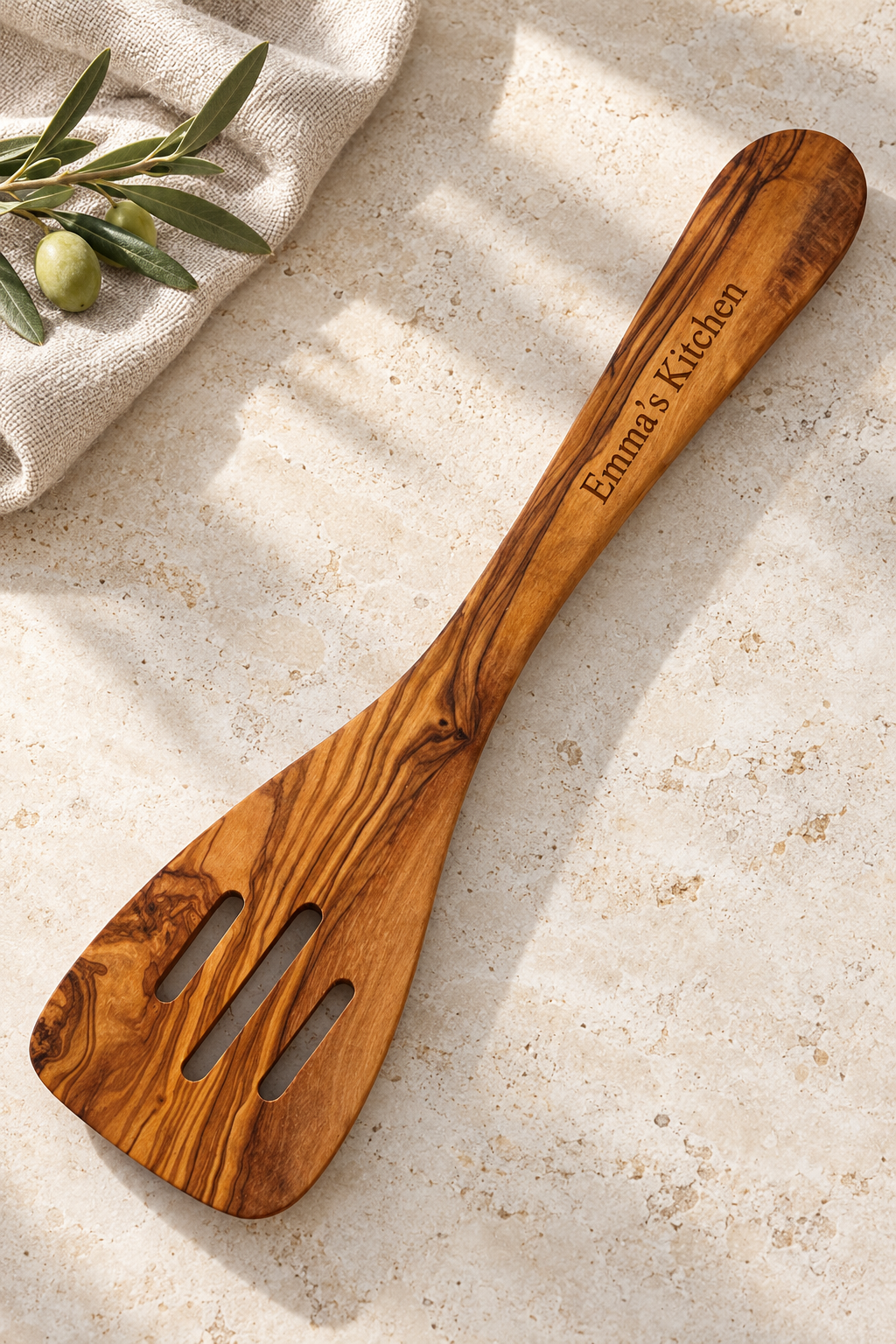 Olive wood slotted spatula engraved “Emma’s Kitchen”, resting on pale stone beside linen and an olive sprig.