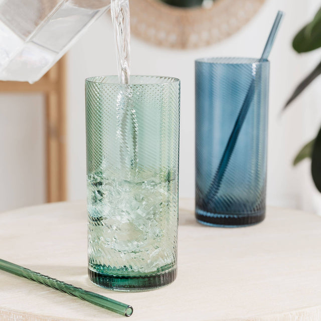 Glass being filled with water on a light surface with another glass and straw in the background.