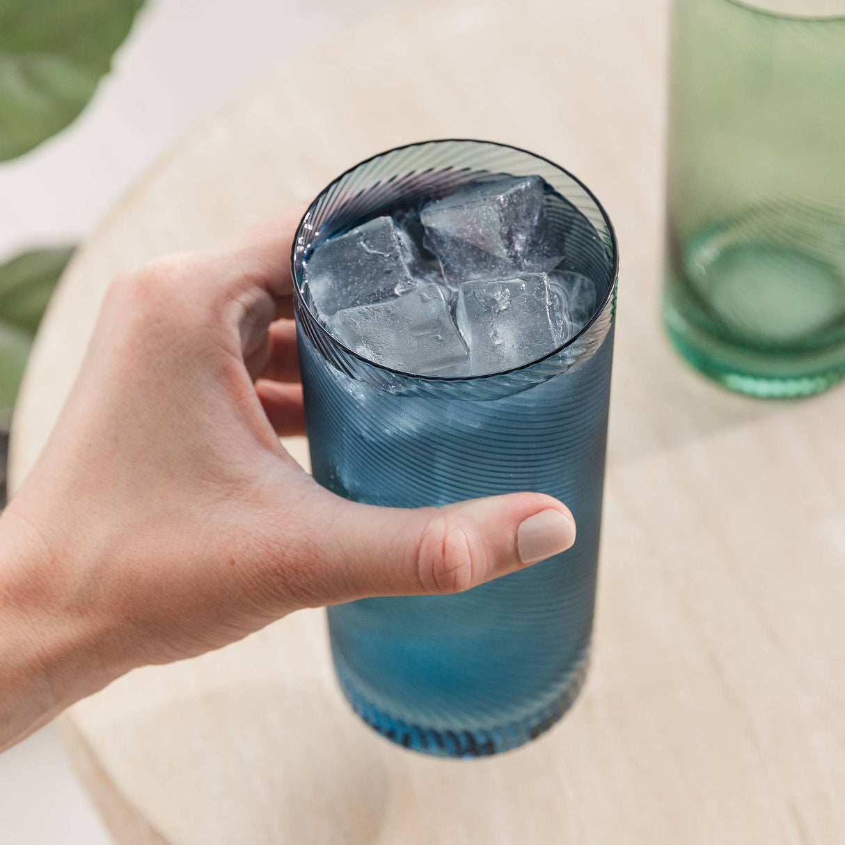 Hand holding a blue glass filled with ice cubes on a light surface.