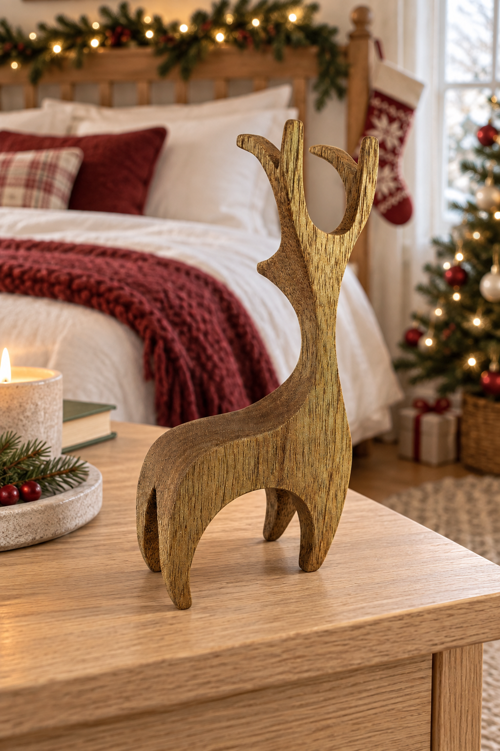 A rustic wooden reindeer figurine stands on a light wood table in a cozy, decorated bedroom setting with a bed, holiday garland, and Christmas tree in the background.