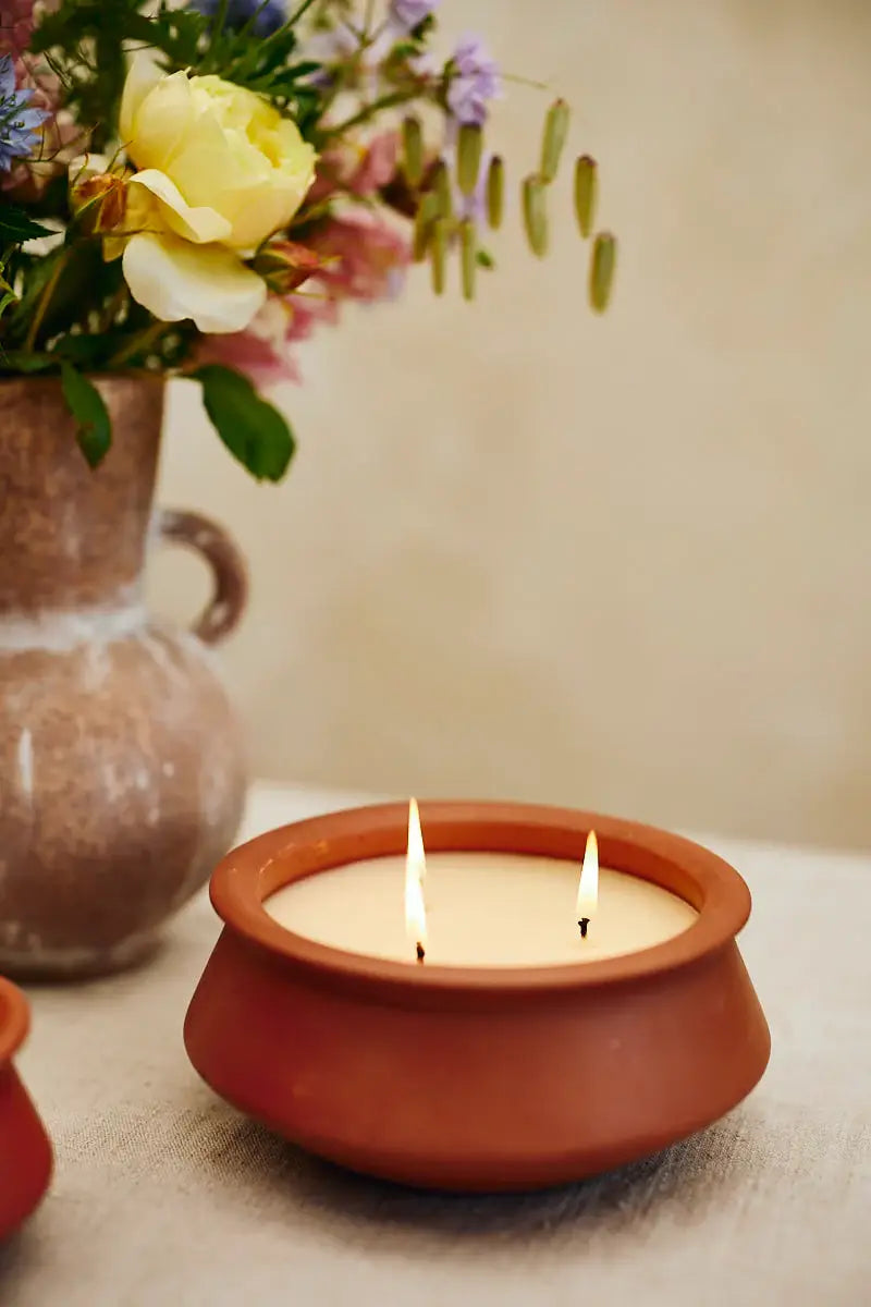 A terracotta clay candle holder with three lit wicks holding a cream-colored candle, set on a light beige surface with a blurred vase of flowers in the background.
Large 3 Wick Terracotta Candle | Lavender & Vanilla