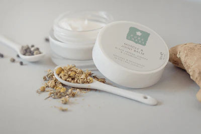Two jars of cream, a spoon with dried herbs, and a piece of ginger on a light surface.