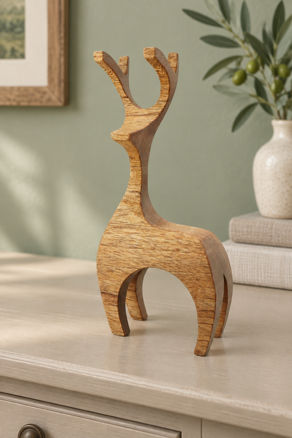 A small sculptural deer figurine made of natural wood with visible grain, displayed on a light-colored cabinet in a styled interior setting with a vase and framed art in the background.