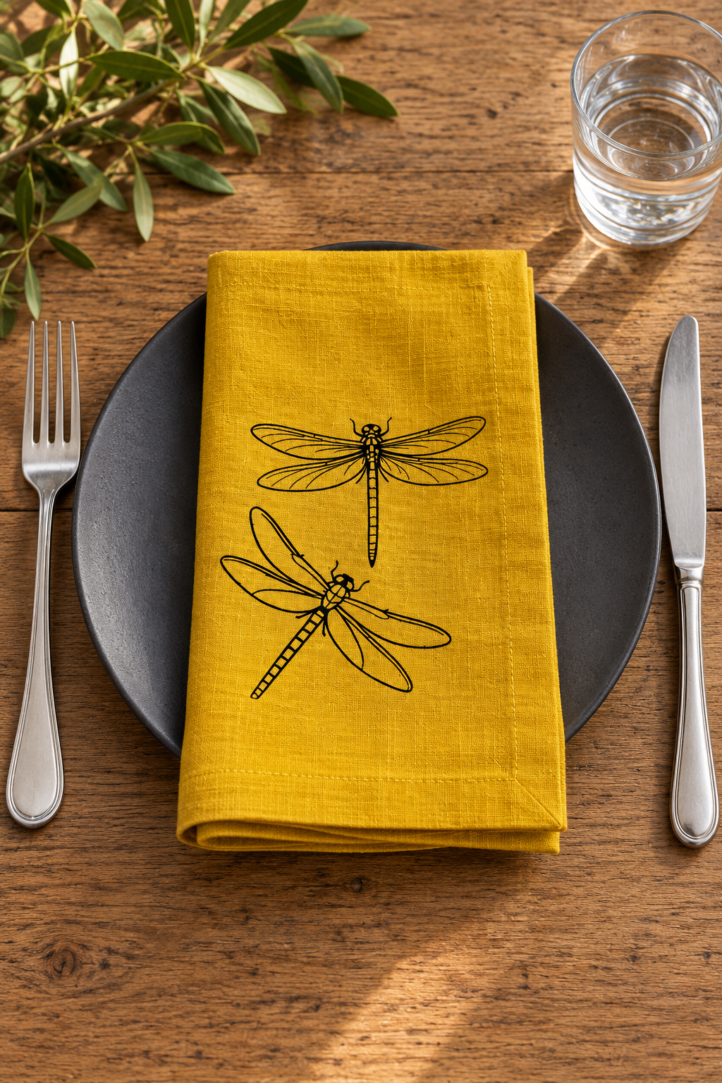 A folded mustard yellow linen napkin with black dragonfly illustrations, displayed on a dark gray plate on a rustic wooden table with silver cutlery and a glass of water.