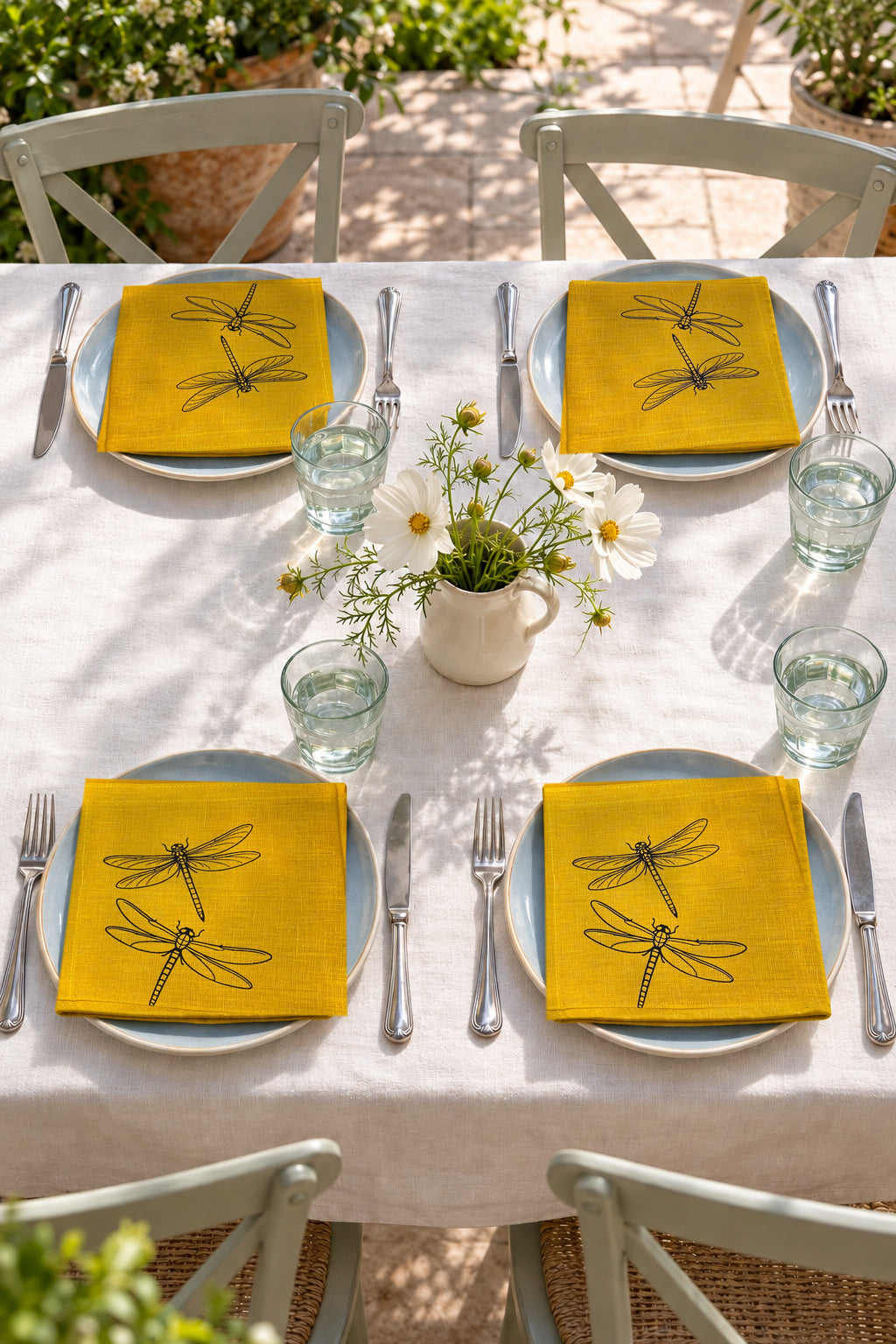 Four yellow linen napkins with black dragonfly are arranged on light blue plates on a white tablecloth in an outdoor garden setting.