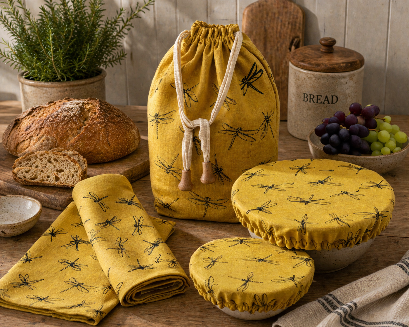 yellow hand printed linen drawstring bread bag, tea towels and bowl covers on a wooden table with bread and grapes.