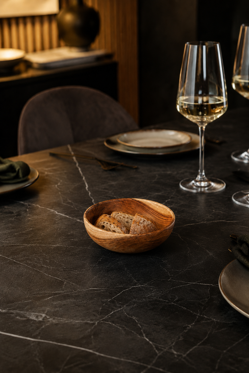 A small round wooden bowl filled with bread sits on a dark marble dining table set with plates, cutlery, and wine glasses in a dimly lit restaurant or dining room.
