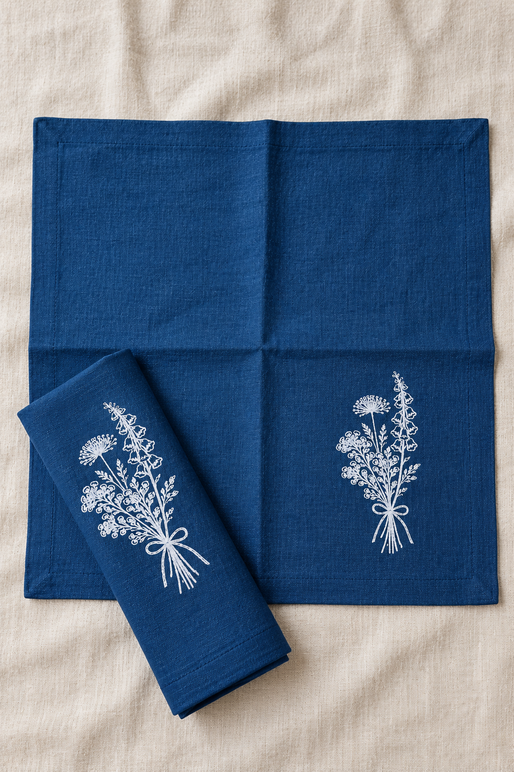 Navy Florilegium linen napkins set on a beige fabric background, featuring white embroidered wildflower bouquets. One napkin is laid flat, and another is folded over to show the embroidered detail.