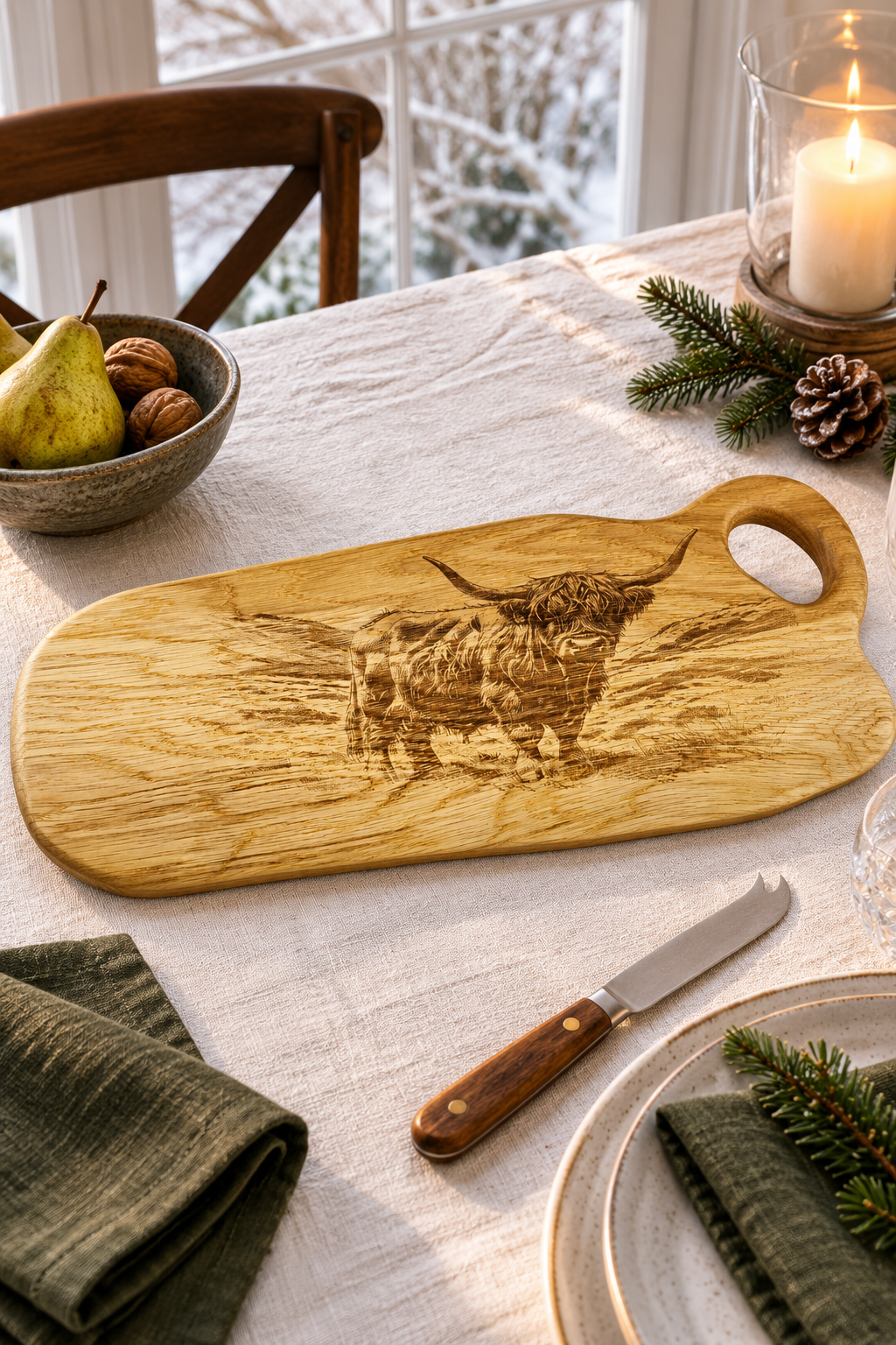 A wooden serving board with a carved Highland cow design, styled on a dining table with a knife, plates, green napkins, a pear, walnuts, pinecones, and a lit candle.