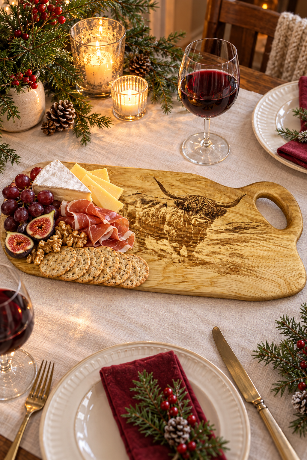 A wooden charcuterie board with an engraved bull design, styled on a dining table with cheese, crackers, grapes, figs, cured meat, wine glasses, candles, and holiday greenery.