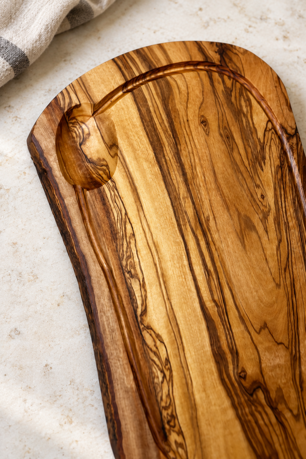 An olive wood serving board with a natural live edge and a carved recessed area, shown on a light textured surface with a folded cloth in the corner.