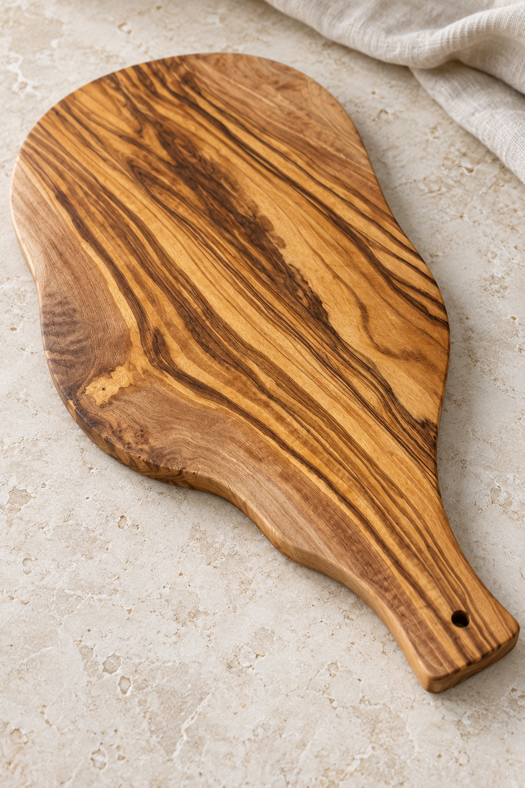 An olive wood serving board with a distinctive dark and light grain pattern, shown on a light beige stone surface with a neutral fabric napkin in the corner.