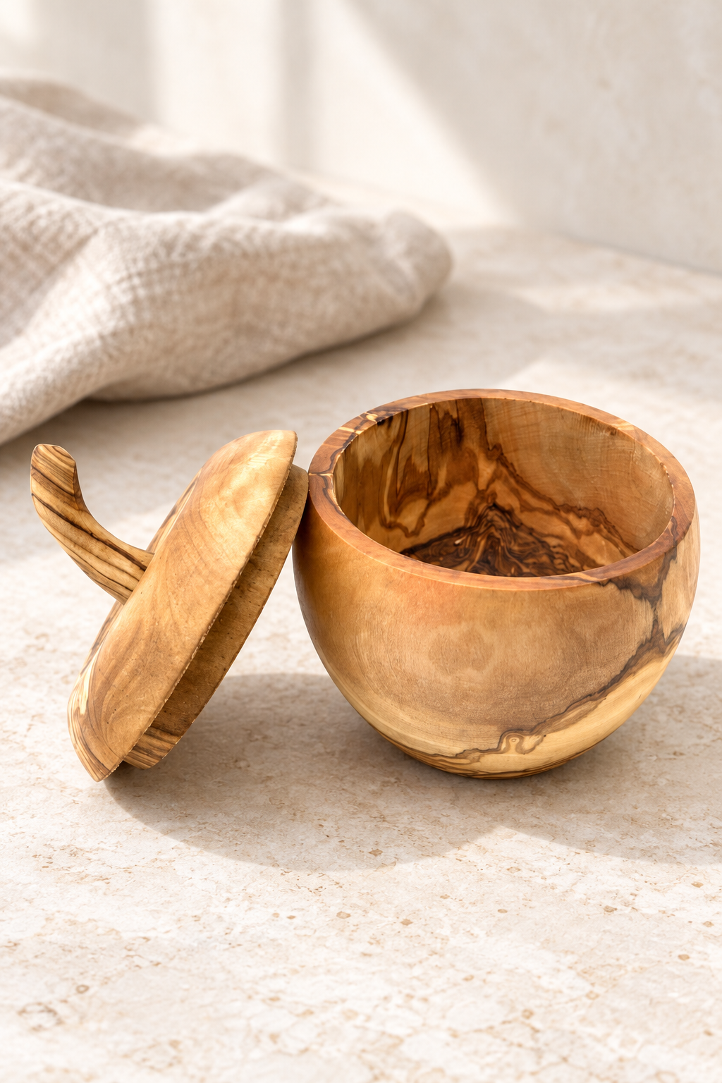 A small handcrafted apple shaped wooden bowl with its lid leaning beside it on a light stone surface. The bowl and lid have a warm honey-brown finish with natural darker wood grain patterns.