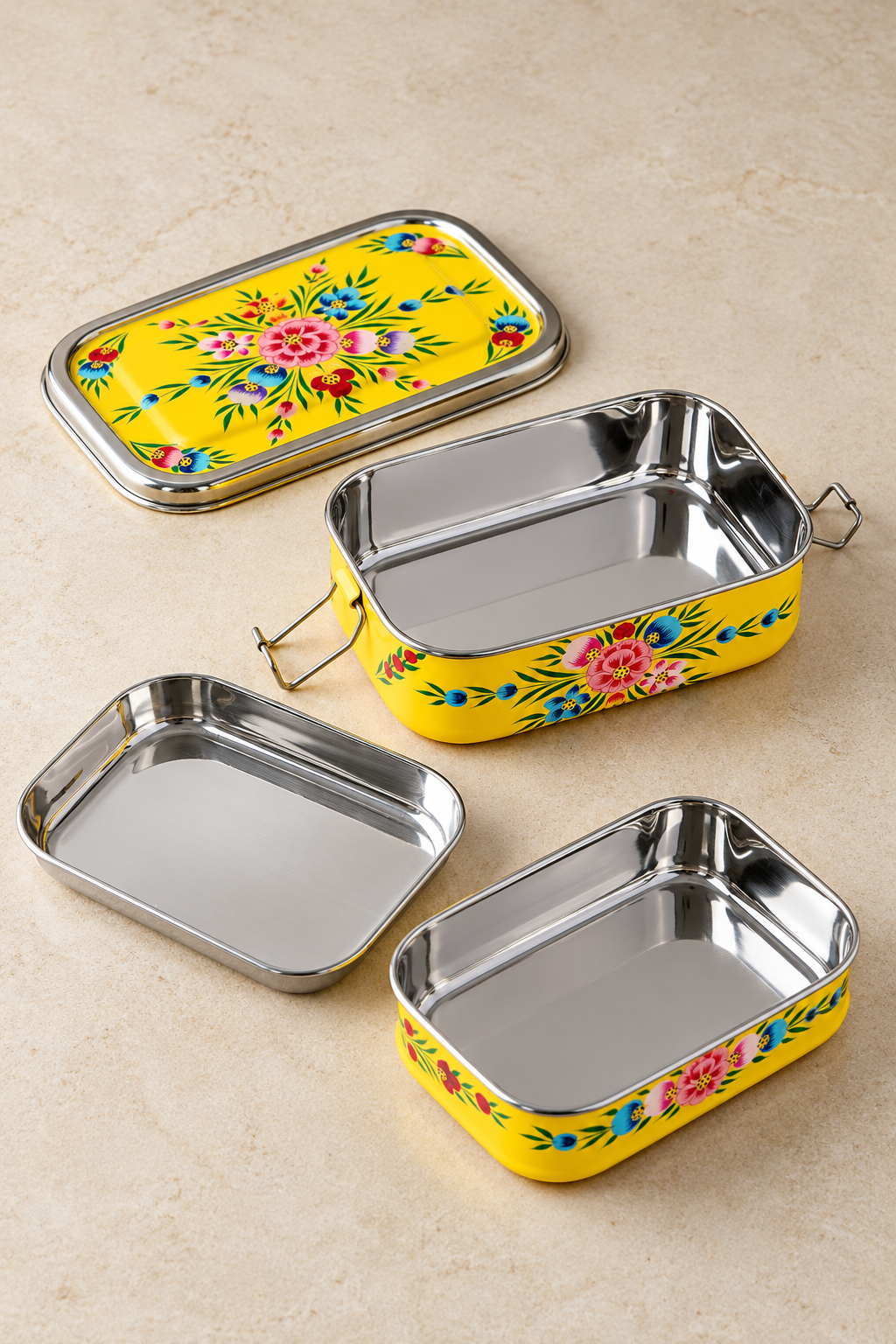 Three rectangular stainless steel lunch boxes with lids, arranged on a beige stone surface. The lids and sides feature a bright yellow background with colorful floral and leaf patterns in pink, blue, green, and red.