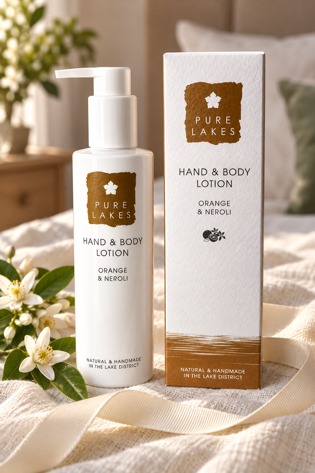 A bottle of Pure Lakes Hand & Body Lotion and its matching outer box, both labeled Orange & Neroli, styled on a cream fabric surface with white flowers and a soft indoor background.