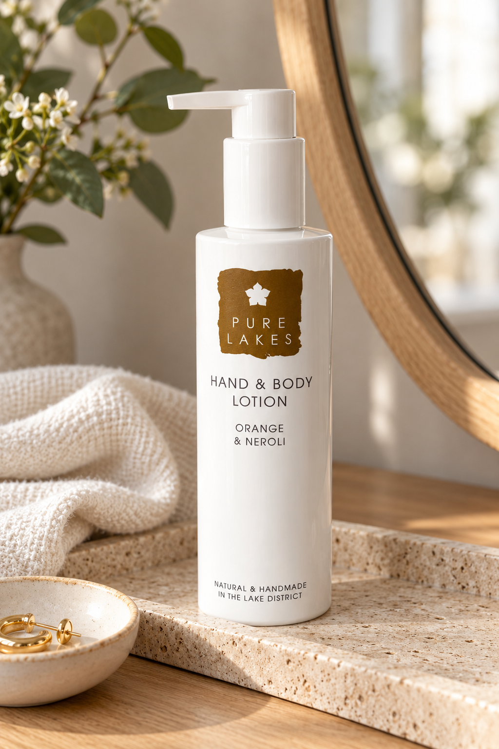 A bottle of Pure Lakes Hand & Body Lotion in Orange & Neroli scent, standing on a speckled stone tray next to a folded cream towel, with a wooden mirror and soft floral decor in the background.