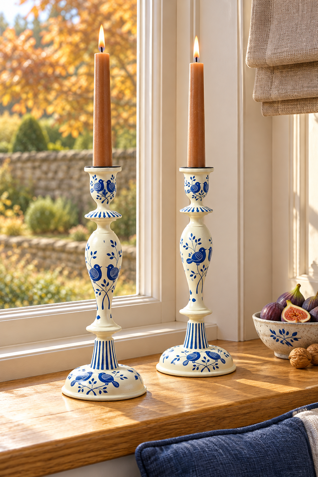 Two tall wood candlesticks with blue floral patterns and lit taper candles, displayed on a wooden windowsill in a bright room.