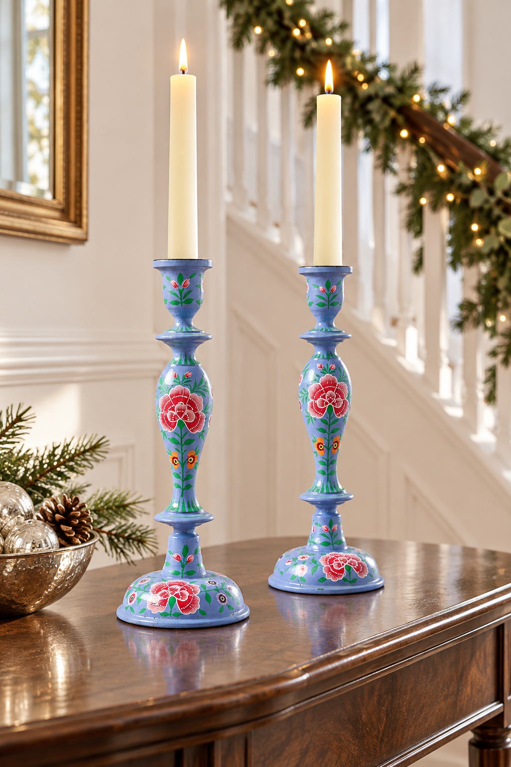 Two tall blue ceramic candlestick holders with floral hand-painted details, each holding a lit white taper candle, displayed on a wooden console table in a decorated indoor staircase setting.