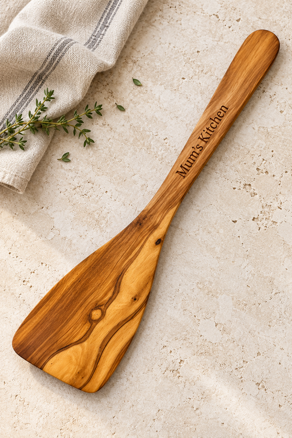 Solid olive wood spatula engraved “Mum’s Kitchen”, resting on pale stone beside striped linen and thyme.