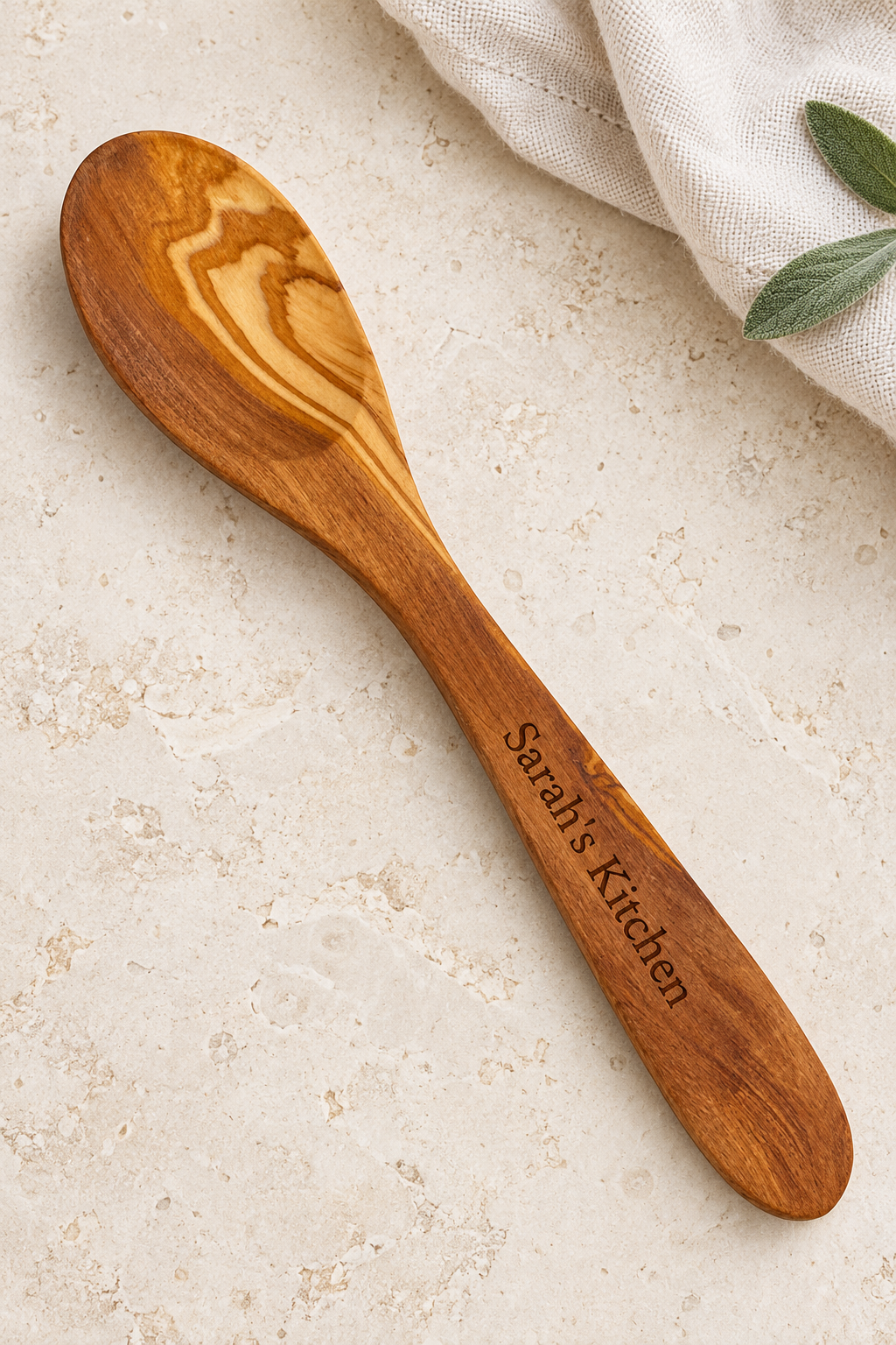 Small olive wood serving spoon with “Sarah’s Kitchen” laser engraved along the handle, displayed on pale stone beside cream linen and sage leaves.