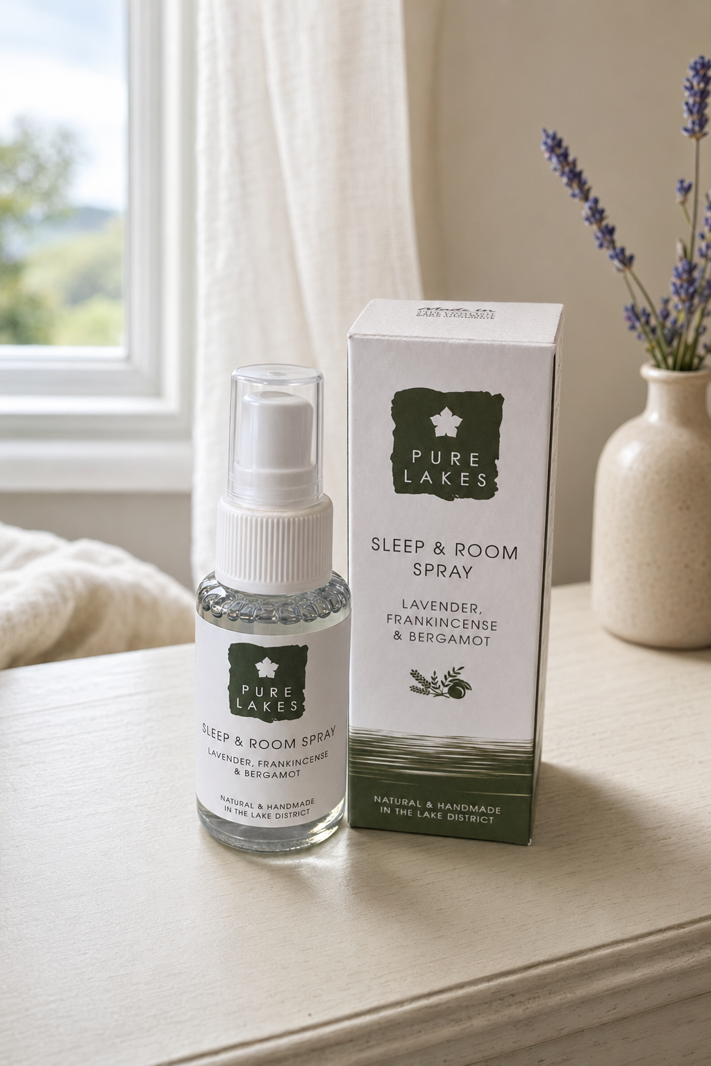 A bottle of Pure Lakes Sleep & Room Spray with its white packaging box, styled on a light wooden table next to a window and a vase of lavender.