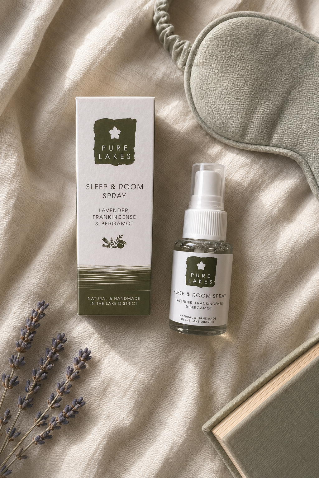 Pure Laces Sleep & Room Spray bottle and its box displayed on a beige linen surface with a gray sleep mask, lavender sprigs, and a book as styled props.