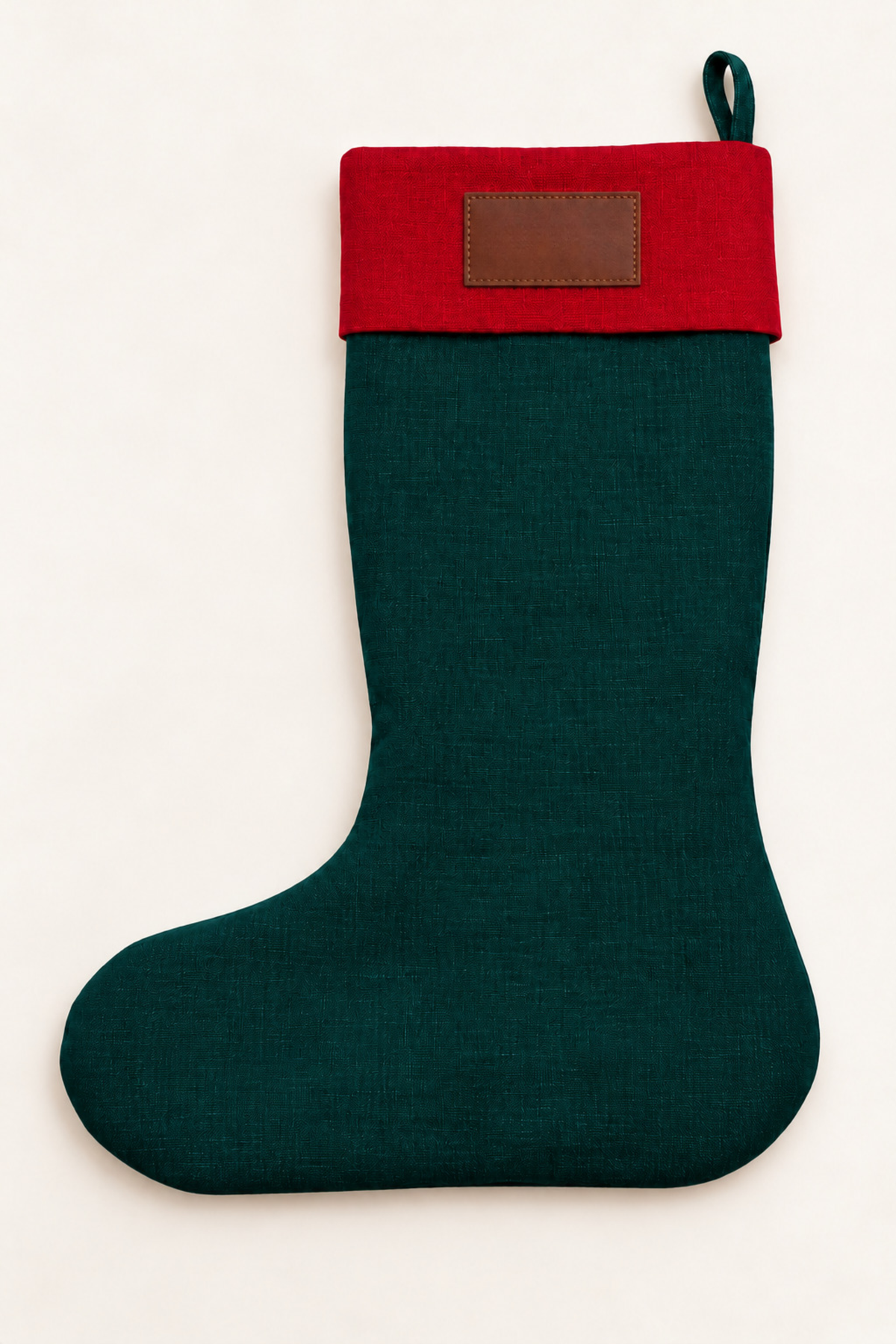 A dark green linen Christmas stocking with a red cuff and a brown leather rectangular patch, shown against a plain off-white background.