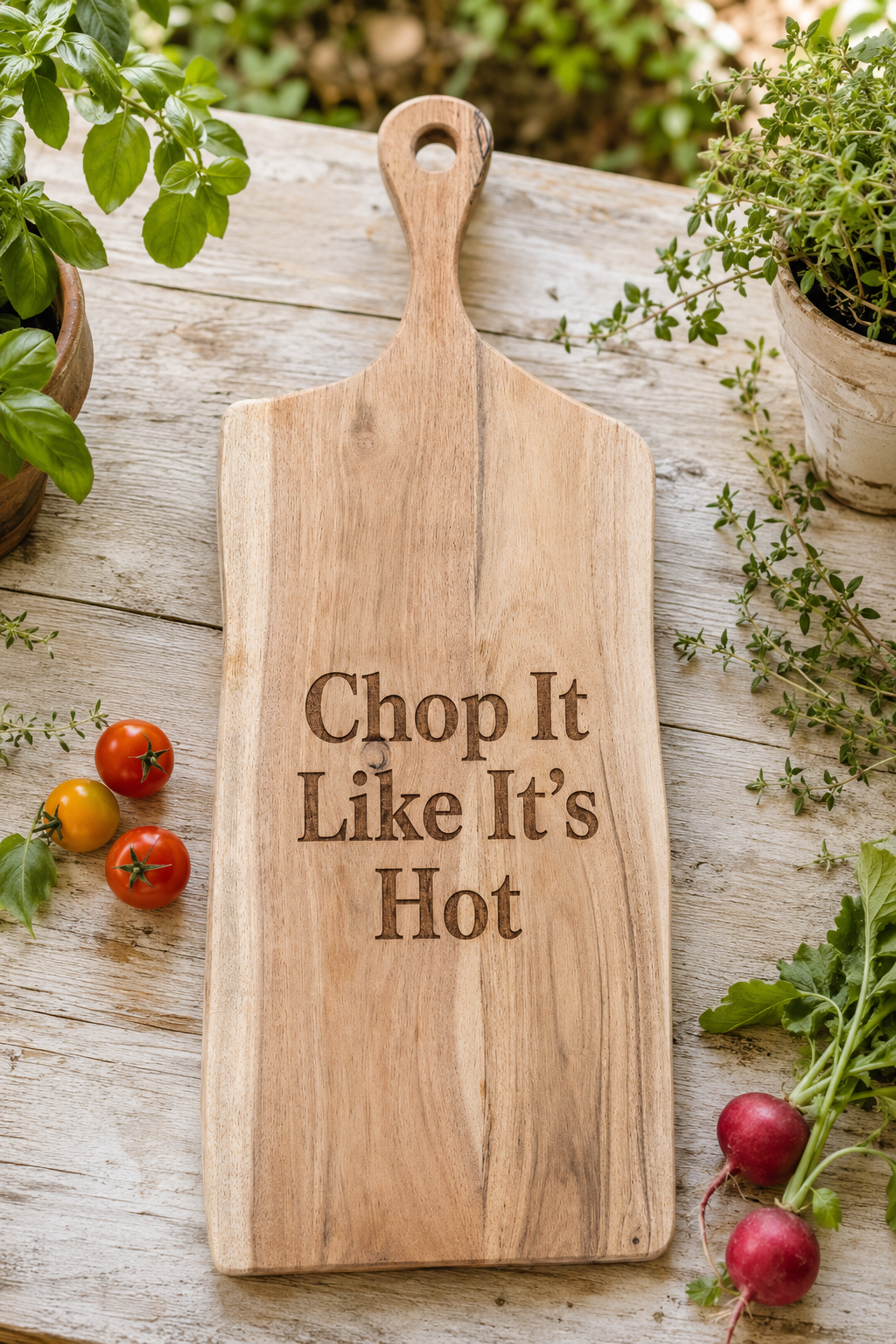 A rectangular wooden cutting board with a handle, engraved with the phrase 'Chop It Like It's Hot' in dark lettering. The board is styled on a weathered wooden table surrounded by fresh green herbs, cherry tomatoes, and radishes.
