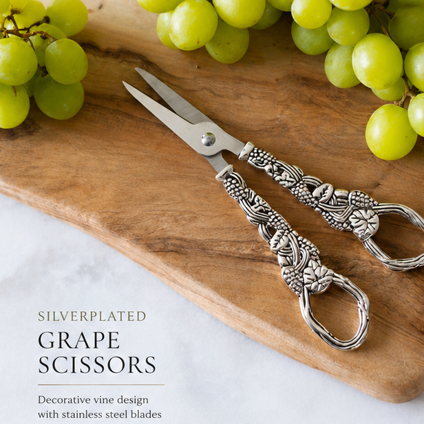 Silver grape scissors with decorative design on a wooden board with grapes and cheese.