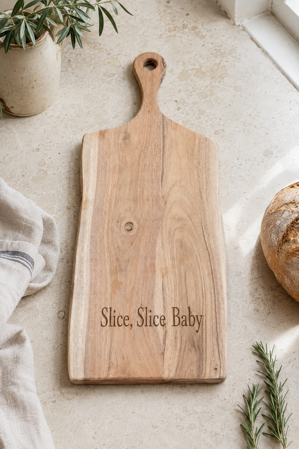 A rectangular wooden serving board with a rounded handle and the engraved phrase 'Slice, Slice Baby' at the bottom, styled on a light stone surface with a cloth, bread, and greenery nearby.