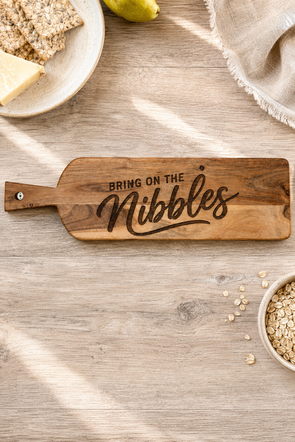 A wooden snack nibble board with the phrase 'BRING ON THE Nibbles' engraved on it, styled on a light wood tabletop with crackers, a pear, oats, and a napkin nearby.