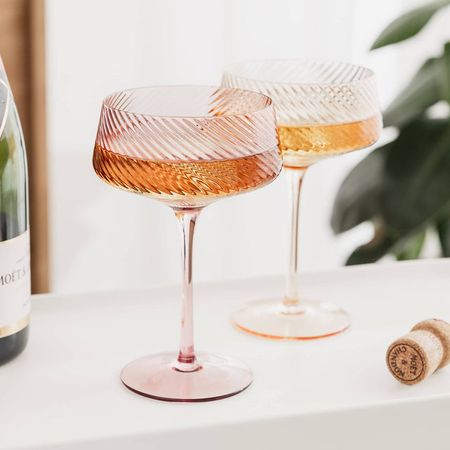Two coupe glasses filled with rose wine on a white surface with a bottle and cork in the background.