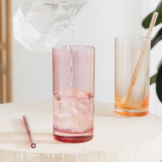 Two tall glasses with textured twist design on a light surface, one pink and one orange, with straws included.