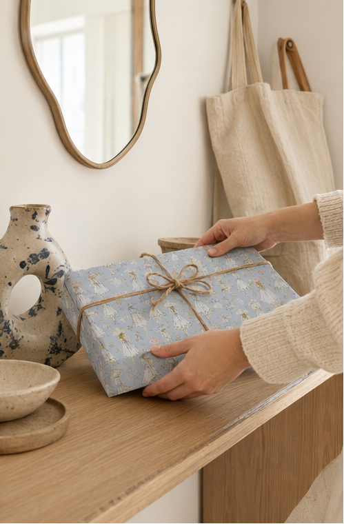 A person's hands are shown holding a gift box wrapped in Vintage Fashion Sketch Wrapping Paper in Pastel Blue and tied with a rustic brown twine bow. The box is placed on a light wood console table in a cozy, neutral-toned room with a speckled vase, a canvas tote bag, and a mirror in the background.