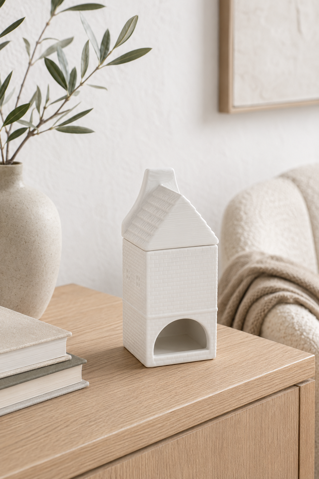 A small White Nordic House Oil Burner & Wax Warmer sits on a light wood side table in a softly styled room. A beige vase with green branches, stacked books, and part of a cream armchair are visible in the background.