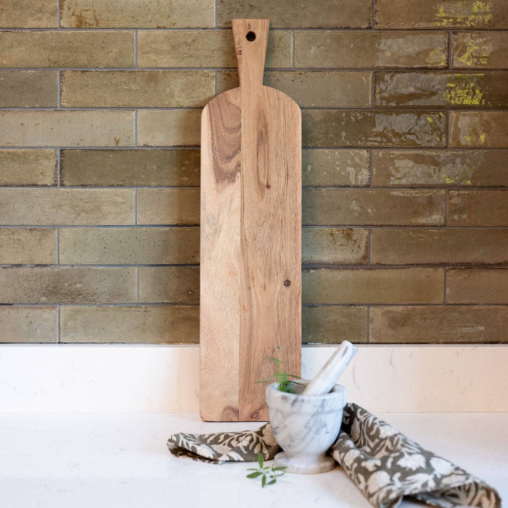 Wooden cutting board on a stone wall with a white surface and kitchen items.