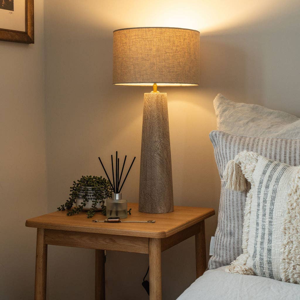Decorative lamp on a wooden table next to a bed with pillows and a blanket.