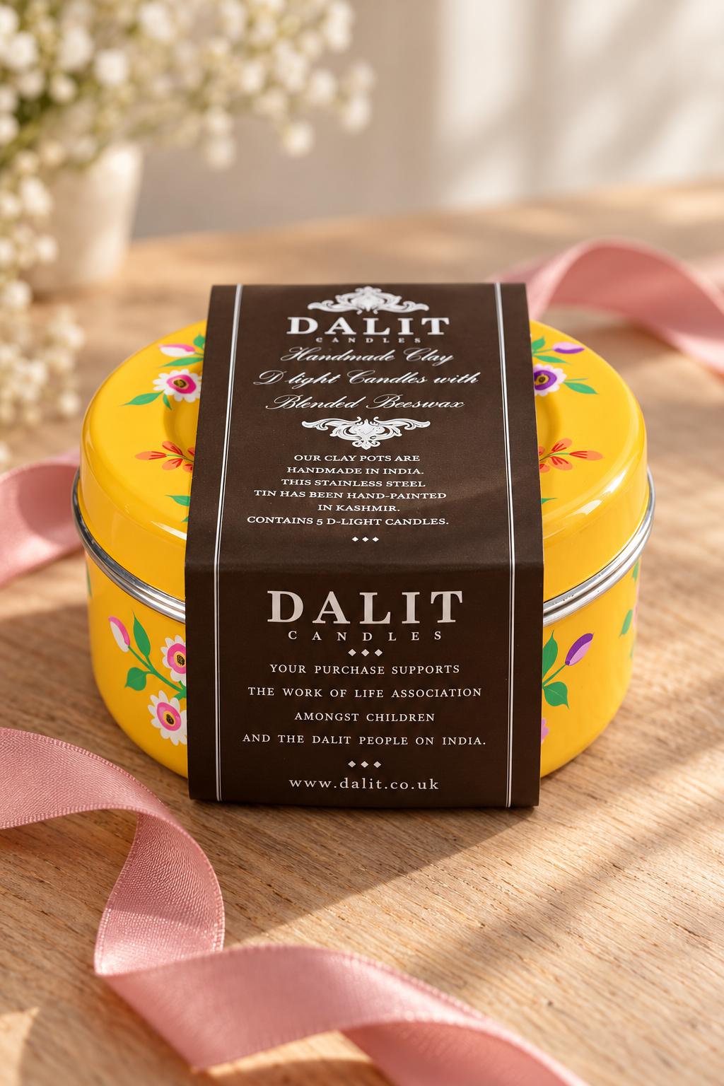 A yellow round tin candle with a dark brown paper label reading DALIT Candles, placed on a wooden surface with pink ribbon and soft floral decor in the background.