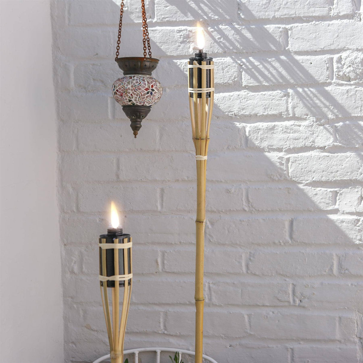 Two lit tiki torches with a hanging lantern against a white brick wall.